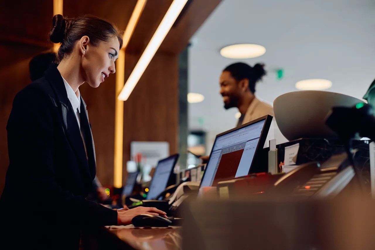 Female receptionist using desktop PC while working at hotel reception desk.